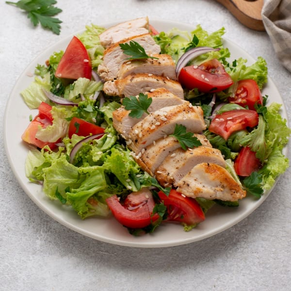 Grilled chicken breast slices with sesame seeds, fresh lettuce, tomatoes, and red onions on a white plate.